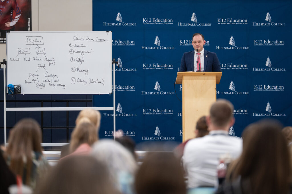A presenter stands beside a whiteboard outlining rhetoric and Cicero’s five canons.